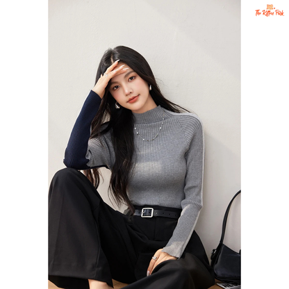 Woman wearing a gray ribbed sweater and black pants sitting against a light gray wall.