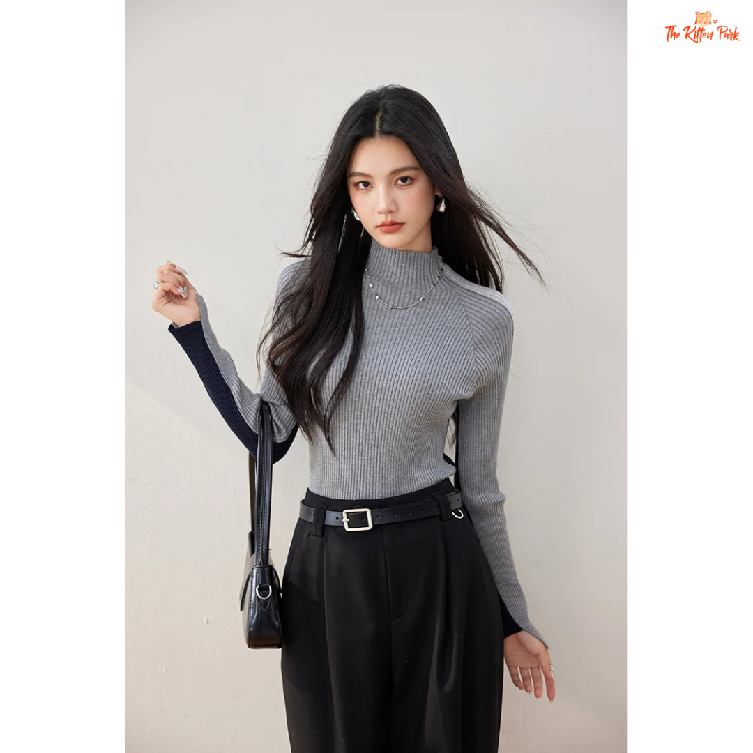 Woman wearing a gray turtleneck and black pants with a black handbag against a plain background