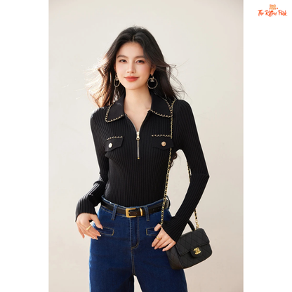 Woman wearing a black ribbed sweater and blue jeans, holding a black handbag against a plain background.