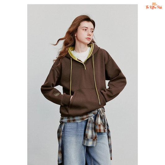 Woman wearing a brown hoodie with yellow trim, plaid shirt, and jeans on a gray background